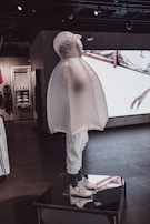 A mannequin dressed in a unique, inflated outerwear piece and white sneakers. The figure is displayed on a platform inside a modern store with dark floors and ambient lighting. Behind it, there are shelves and a large screen showing abstract visuals.