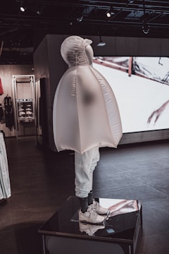 A mannequin dressed in a unique, inflated outerwear piece and white sneakers. The figure is displayed on a platform inside a modern store with dark floors and ambient lighting. Behind it, there are shelves and a large screen showing abstract visuals.