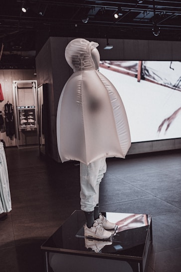 A mannequin dressed in a unique, inflated outerwear piece and white sneakers. The figure is displayed on a platform inside a modern store with dark floors and ambient lighting. Behind it, there are shelves and a large screen showing abstract visuals.
