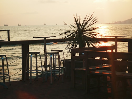 A serene seaside setting with a wooden deck featuring bar stools and tables. A palm plant is silhouetted against the golden hues of a sunset reflecting on the calm sea. Ships are visible on the horizon under a softly cloudy sky.