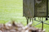 A weathered electrical box stands on a metal frame in an outdoor setting, with visible signs of moss and dirt covering its surface. Several cables extend from the bottom of the unit. The background is a blurred expanse of greenery, possibly grass.
