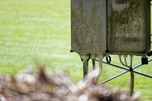 A weathered electrical box stands on a metal frame in an outdoor setting, with visible signs of moss and dirt covering its surface. Several cables extend from the bottom of the unit. The background is a blurred expanse of greenery, possibly grass.