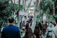 A group of people gathered outdoors for a ceremony, likely a wedding, surrounded by lush greenery and trees. A man stands at the front, seemingly speaking to the audience, with an arch and drapery behind him. Guests are seated on wooden chairs, facing the speaker.