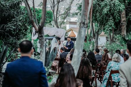 A group of people gathered outdoors for a ceremony, likely a wedding, surrounded by lush greenery and trees. A man stands at the front, seemingly speaking to the audience, with an arch and drapery behind him. Guests are seated on wooden chairs, facing the speaker.