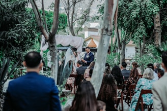 A group of people gathered outdoors for a ceremony, likely a wedding, surrounded by lush greenery and trees. A man stands at the front, seemingly speaking to the audience, with an arch and drapery behind him. Guests are seated on wooden chairs, facing the speaker.