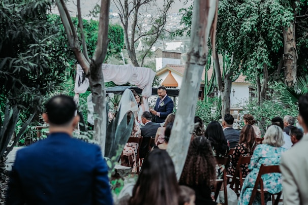 A group of people gathered outdoors for a ceremony, likely a wedding, surrounded by lush greenery and trees. A man stands at the front, seemingly speaking to the audience, with an arch and drapery behind him. Guests are seated on wooden chairs, facing the speaker.