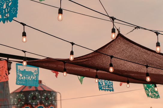 Strings of light bulbs hang across a patio space with a brown fabric canopy. Colorful papel picado banners with intricate designs are suspended in the air, while an ornately painted structure with circular patterns is visible in the background under a warm, dusky sky.