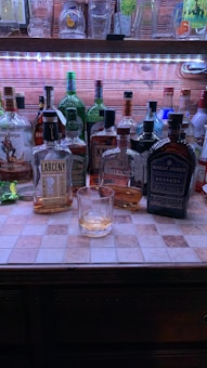 A collection of various liquor bottles is arranged on a tiled countertop with a wooden background. A filled glass is placed in front of the bottles, which include brands like Larceny, Jefferson's, and Great Jones Bourbon. Above, empty glasses and mugs are displayed on a shelf with a LED light.