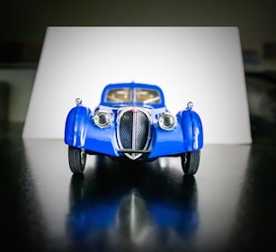 A miniature model of a vintage blue car is placed against a neutral background. The car exhibits a classic design with prominent round headlights, a distinctive front grille, and detailed curved fenders. The wheels and other features are accurately scaled to mimic the original vehicle.