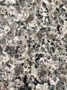 A close-up view of a granite surface with a coarse, speckled pattern. The stone features a mix of black, white, and gray crystals with some pinkish areas, giving it a textured and natural appearance.