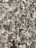 A close-up view of a granite surface with a coarse, speckled pattern. The stone features a mix of black, white, and gray crystals with some pinkish areas, giving it a textured and natural appearance.