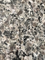 A close-up view of a granite surface with a coarse, speckled pattern. The stone features a mix of black, white, and gray crystals with some pinkish areas, giving it a textured and natural appearance.