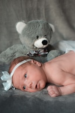 A newborn baby lies on a soft, grey surface with a large grey teddy bear positioned behind. The baby wears a white headband with a bow, displaying an alert and curious expression.