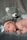 A newborn baby lies on a soft, grey surface with a large grey teddy bear positioned behind. The baby wears a white headband with a bow, displaying an alert and curious expression.