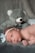A newborn baby lies on a soft, grey surface with a large grey teddy bear positioned behind. The baby wears a white headband with a bow, displaying an alert and curious expression.