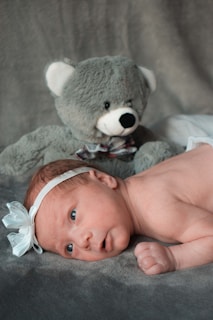 A newborn baby lies on a soft, grey surface with a large grey teddy bear positioned behind. The baby wears a white headband with a bow, displaying an alert and curious expression.