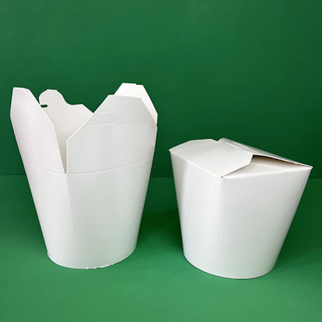 A stack of white and black to go boxes neatly arranged on a wooden table with green plants in the background.