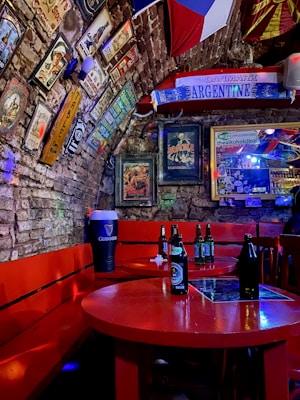 A cozy bar interior with brick walls adorned with various vintage posters and advertisements. Flags and decorative items are displayed, and there is a vibrant red seating area with wooden benches and round tables. Beer bottles are placed on the tables, and a large Guinness-branded container is visible.