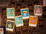 A collection of trading cards from a game called 'Toons and Towers' is spread out on a wooden surface. Each card features a colorful cartoon character and various stats like class, attack, and defense. The characters include a bird, a mushroom, a pig, a chameleon, a cat, and a large bird-like creature.