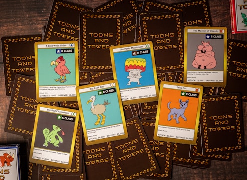 A collection of trading cards from a game called 'Toons and Towers' is spread out on a wooden surface. Each card features a colorful cartoon character and various stats like class, attack, and defense. The characters include a bird, a mushroom, a pig, a chameleon, a cat, and a large bird-like creature.