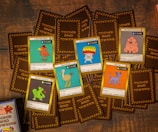 A collection of trading cards is laid out on a wooden surface. The cards feature cartoonish illustrations of various creatures with humorous names like 'A Bird With Slides,' 'The Party Mushroom,' and 'Izita Cat.' The brown backing of several cards shows the logo 'Toons and Towers.' Each card includes stats like 'Attack' and 'Defense' and a playful description.