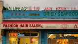 A storefront with multiple businesses, including a fashion hair salon, an insurance agency, and a shop selling ginseng and dried seafood. The facade is colorful with signs in both English and another language. Neon lights and an open sign are visible in the salon window.