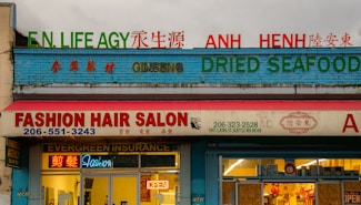 A storefront with multiple businesses, including a fashion hair salon, an insurance agency, and a shop selling ginseng and dried seafood. The facade is colorful with signs in both English and another language. Neon lights and an open sign are visible in the salon window.