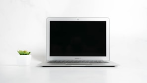 A laptop with a closed screen sits on a clean, white surface. To the left of the laptop is a small potted succulent plant in a white planter. The background is minimalistic and white, emphasizing the simplicity and cleanliness of the scene.