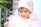 A young child wearing a pink knit hat with floral decorations and fluffy pom-poms. The child is dressed in a pastel-colored outfit that includes a tulle detail. Bright natural lighting creates a soft and gentle ambiance, with greenery and a blurred background providing a peaceful setting.