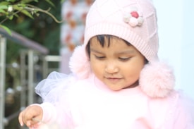 A young child wearing a pink knit hat with floral decorations and fluffy pom-poms. The child is dressed in a pastel-colored outfit that includes a tulle detail. Bright natural lighting creates a soft and gentle ambiance, with greenery and a blurred background providing a peaceful setting.