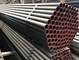 A stack of metal pipes, neatly arranged and secured with straps. The pipes have a uniform cylindrical shape and are aligned horizontally across the image. The visible ends of the pipes show a reddish tint, contrasting with the darker metal body.