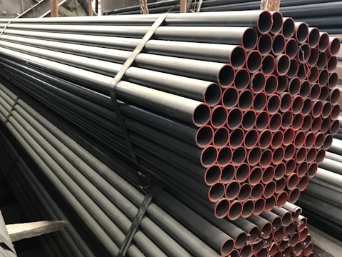 A stack of metal pipes, neatly arranged and secured with straps. The pipes have a uniform cylindrical shape and are aligned horizontally across the image. The visible ends of the pipes show a reddish tint, contrasting with the darker metal body.