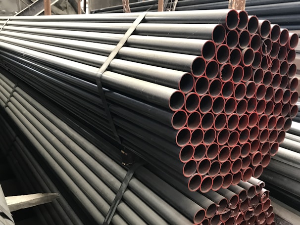 A stack of metal pipes, neatly arranged and secured with straps. The pipes have a uniform cylindrical shape and are aligned horizontally across the image. The visible ends of the pipes show a reddish tint, contrasting with the darker metal body.