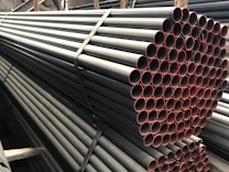 A stack of metal pipes, neatly arranged and secured with straps. The pipes have a uniform cylindrical shape and are aligned horizontally across the image. The visible ends of the pipes show a reddish tint, contrasting with the darker metal body.