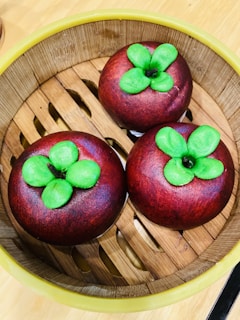 Three round, dark red pastries with vibrant green leaves on top are placed in a bamboo steamer basket. The basket has a natural light brown color with a woven design, surrounded by a yellow rim. The pastries mimic the appearance of mangosteen fruit.