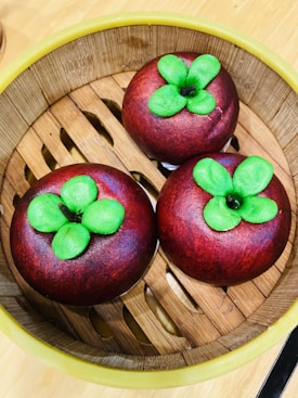 Three round, dark red pastries with vibrant green leaves on top are placed in a bamboo steamer basket. The basket has a natural light brown color with a woven design, surrounded by a yellow rim. The pastries mimic the appearance of mangosteen fruit.