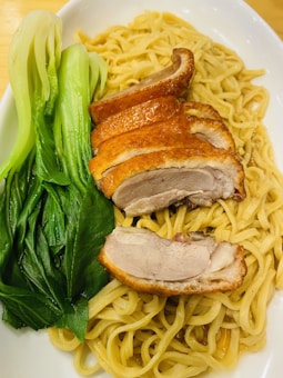 A plate of noodles topped with sliced roasted duck breast and accompanied by steamed green bok choy. The noodles appear lightly coated in a sauce, and the dish is presented on a white plate.