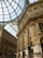 A grand, ornate shopping arcade with glass domed roof and intricately designed facades. The interior architecture features detailed stone carvings, arched windows, and prominent brand stores.