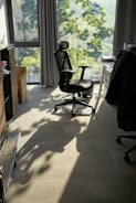 A modern office chair is positioned near a large window with sunlight streaming in, casting shadows on the carpeted floor. The room features a desk with papers and a dark jacket draped over another chair. Green leaves outside the window suggest a tree-lined view.
