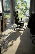 A modern office chair is positioned near a large window with sunlight streaming in, casting shadows on the carpeted floor. The room features a desk with papers and a dark jacket draped over another chair. Green leaves outside the window suggest a tree-lined view.