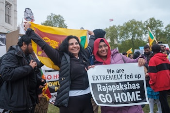 A group of people are gathered at a protest. Two women are smiling and holding a flag and a sign that reads 'We are EXTREMELY fed up Rajapakshas GO HOME.' Other protesters are visible in the background, holding signs and flags. The setting appears to be outdoors, with trees and an overcast sky.