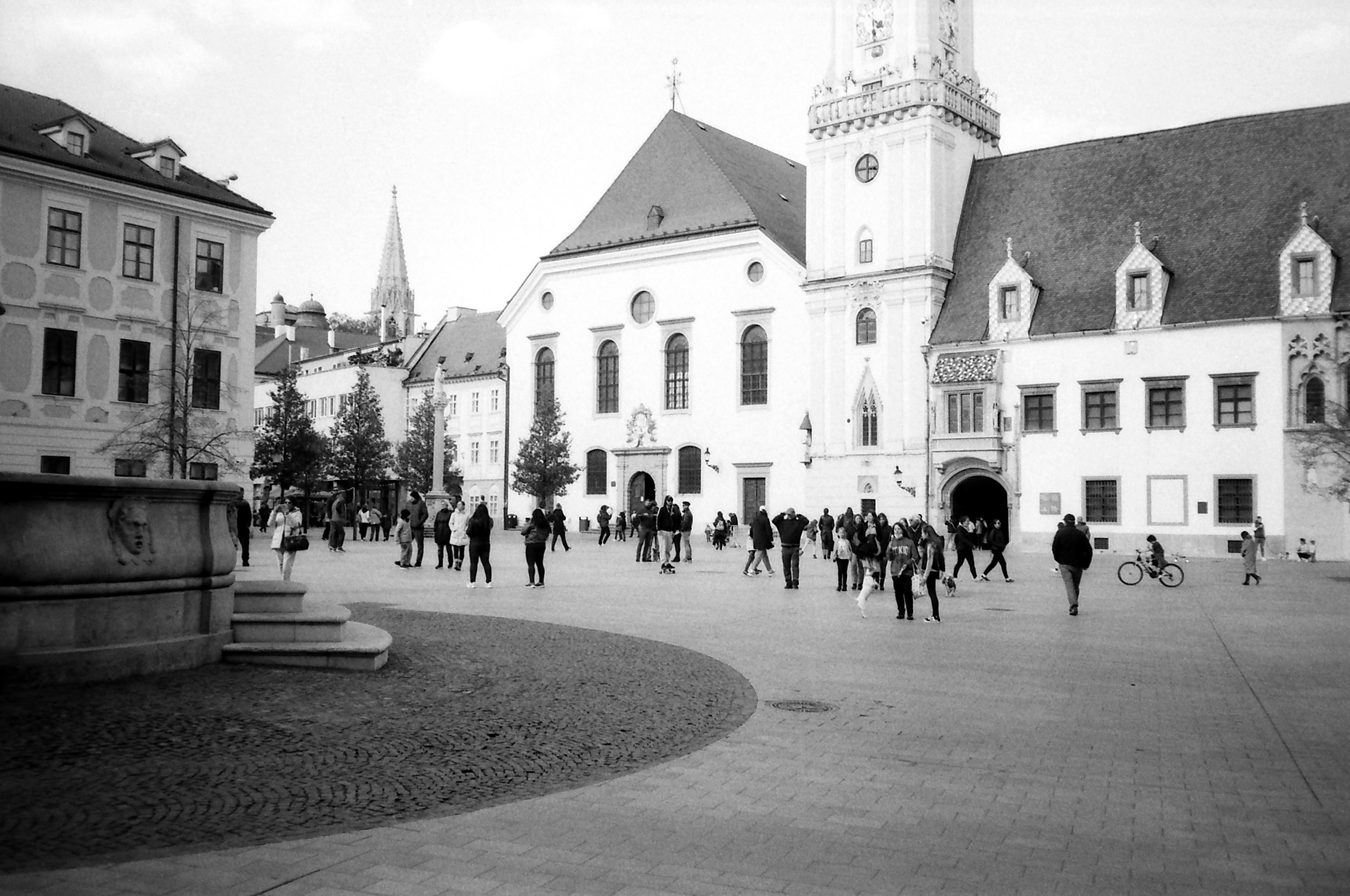 a group of people walking in front of a building, Main Square, Bratislava, Slovakia. Easter Sunday, 2022. Olympus XA on Agfa APX 100 B&W film.