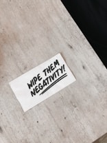 A white napkin with bold black lettering that reads 'WIPE THEM NEGATIVITY!' is placed on a light gray, textured surface.
