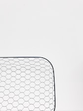 A close-up view of a hexagonal wire mesh, likely made of metal, against a white background. The mesh forms a curved rectangular shape, consisting of evenly spaced hexagonal patterns.