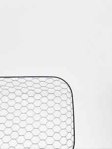 A close-up view of a hexagonal wire mesh, likely made of metal, against a white background. The mesh forms a curved rectangular shape, consisting of evenly spaced hexagonal patterns.