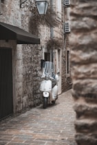 A white scooter is parked on a narrow cobblestone alley surrounded by rustic stone buildings. A vintage-style street lamp hangs above, and there are small, recessed windows with vines.