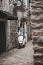 A white scooter is parked on a narrow cobblestone alley surrounded by rustic stone buildings. A vintage-style street lamp hangs above, and there are small, recessed windows with vines.