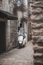 A white scooter is parked on a narrow cobblestone alley surrounded by rustic stone buildings. A vintage-style street lamp hangs above, and there are small, recessed windows with vines.