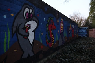A vibrant mural painted on a wall featuring cartoon-style aquatic creatures. The mural includes a large grey fish holding a drink, a red sea dragon with green scales, and a purple creature wearing a crown. Smaller fish and marine plants are scattered throughout the scene, set against a deep blue ocean background.