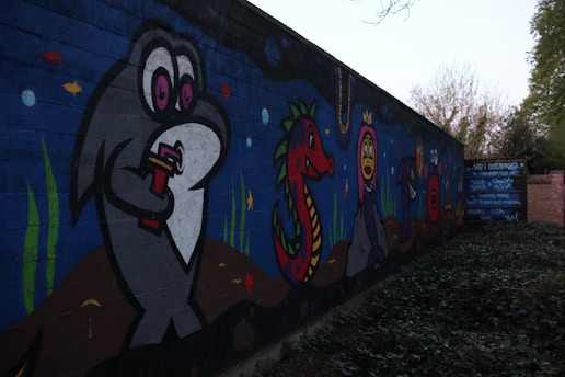 A vibrant mural painted on a wall featuring cartoon-style aquatic creatures. The mural includes a large grey fish holding a drink, a red sea dragon with green scales, and a purple creature wearing a crown. Smaller fish and marine plants are scattered throughout the scene, set against a deep blue ocean background.